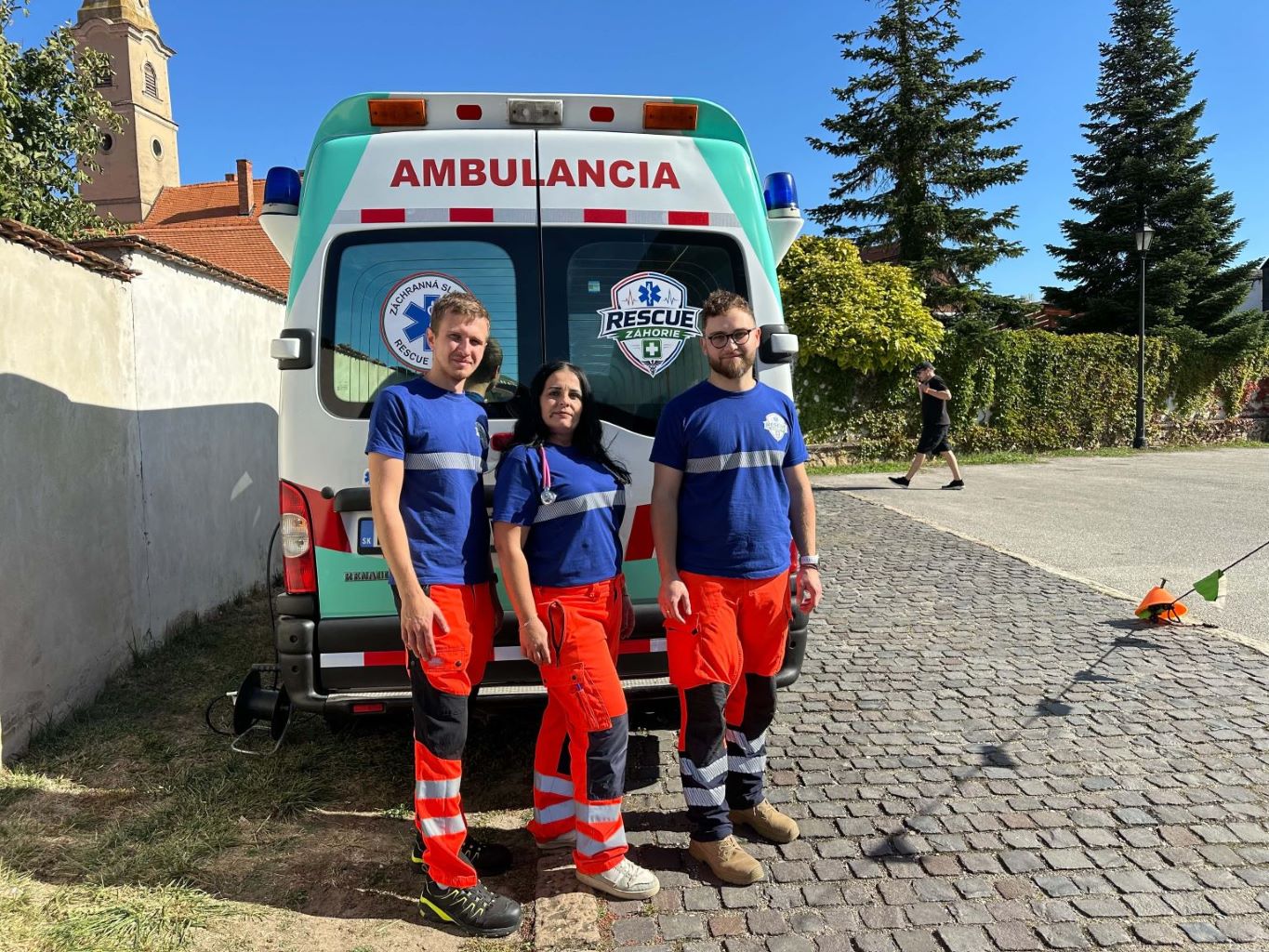 Záchranári v modrých tričkách a oranžových nohaviciach stoja pred sanitkou na ulici.